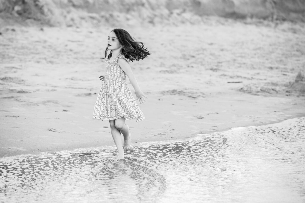 A young girl in a patterned dress runs joyfully along the edge of the water at a sandy beach. The image is black and white, capturing her movement and the gentle waves at her feet. Vero Beach Photoshoot