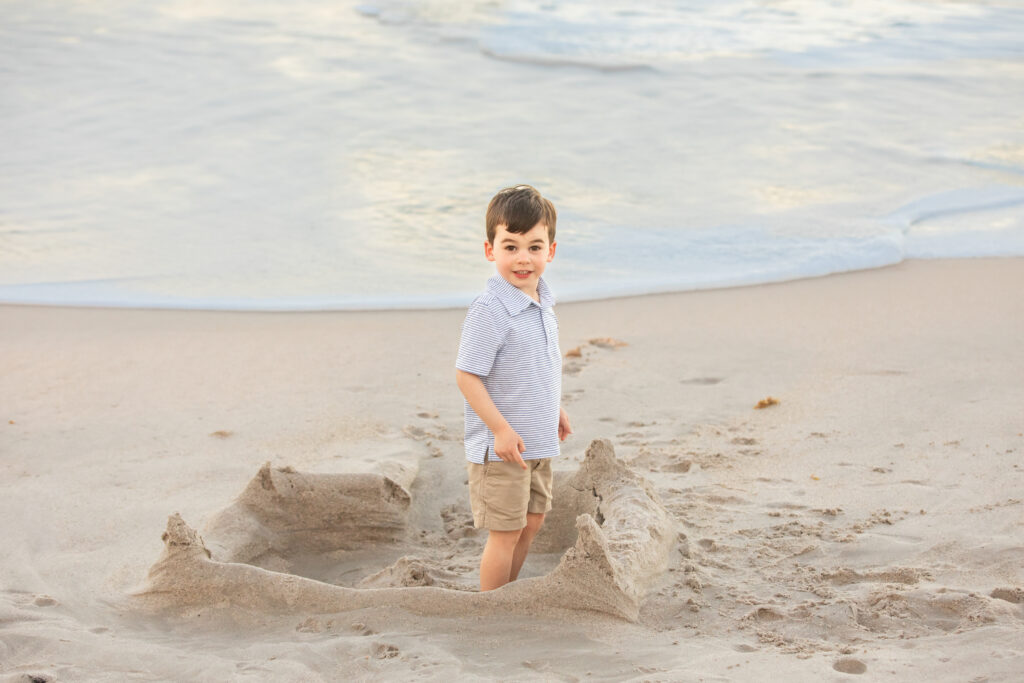 A young boy stands smiling inside a sandcastle on a sandy beach, with gentle ocean waves in the background. He is wearing a striped shirt and khaki shorts. Vero Beach Photoshoot