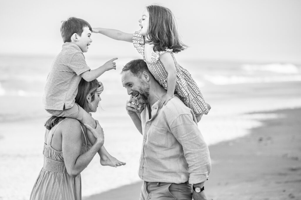 A black and white photo of a smiling family at the beach; a woman carries a young boy on her shoulders while a man carries a young girl on his, with the children reaching out playfully toward each other. Vero Beach Photoshoot