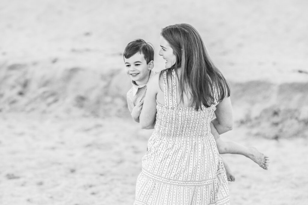 A woman in a patterned dress carries a smiling young boy on a sandy beach. Both appear happy, and the scene is relaxed and playful. The image is in black and white. Vero Beach Photoshoot