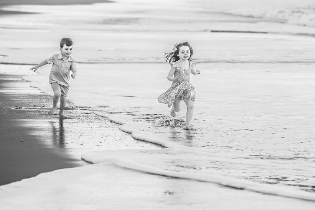 Two young children run and play barefoot along the shoreline, splashing through shallow waves on a sandy beach. The image is in black and white. Vero Beach Photoshoot