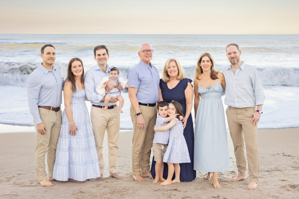Three generations of a family stand barefoot on a beach at sunset, smiling at the camera. Two young children hug in front, while the adults and a baby stand behind, dressed in coordinated blue and white outfits. Waves roll in the background. Vero Beach Photoshoot