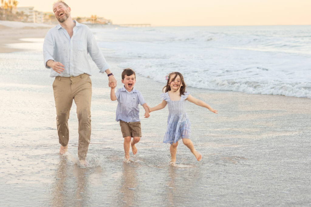 A man and two young children joyfully run barefoot along the shoreline, holding hands, with gentle waves and a sandy beach in the background. Vero Beach Photoshoot