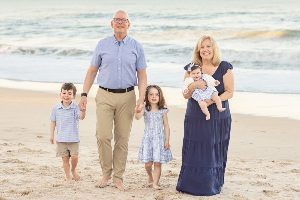 An older man and woman stand barefoot on a beach with three young children, holding hands. The boy and girl are standing, while the woman holds a baby. They are all smiling, with waves and sand in the background. Vero Beach Photoshoot
