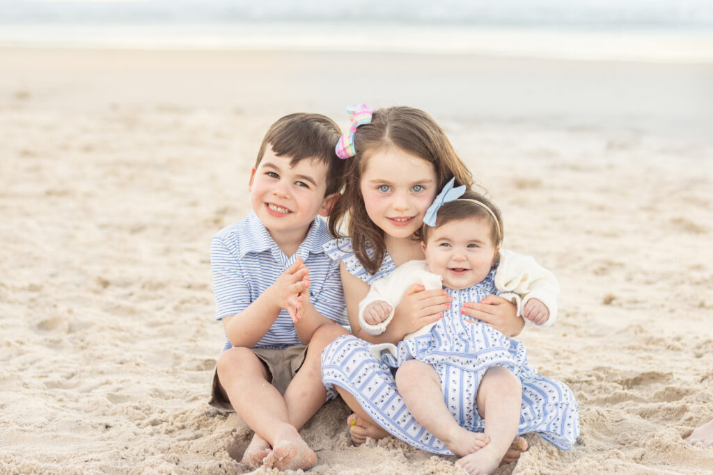 Three smiling children sit together on a sandy beach, with an older girl in the center holding a baby, and a young boy sitting beside them. All are dressed in light, summery clothes. The ocean is in the background. Vero Beach Photoshoot