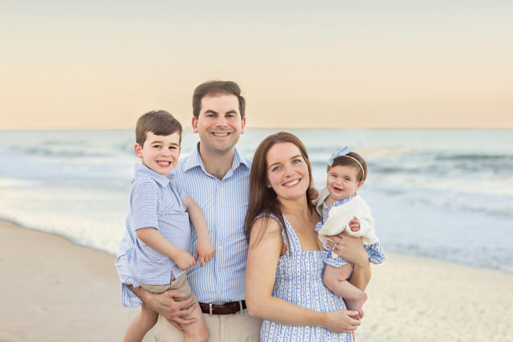 A smiling family of four poses on a beach at sunset. The father holds a young boy, while the mother holds a baby girl. The ocean and sandy shore are visible in the background. All are dressed in light-colored, summery outfits. Vero Beach Photoshoot