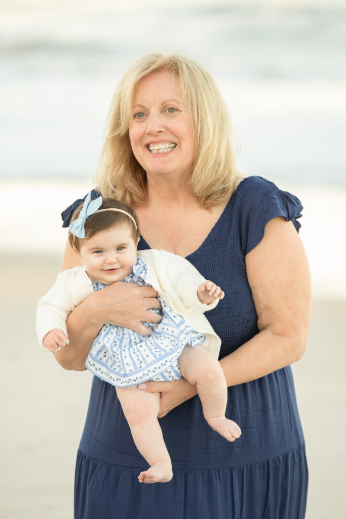A smiling woman with blonde hair, wearing a navy blue dress, holds a baby dressed in a blue-and-white outfit with a bow headband. They are standing outdoors, likely on a beach, with a blurred background. Vero Beach Photoshoot