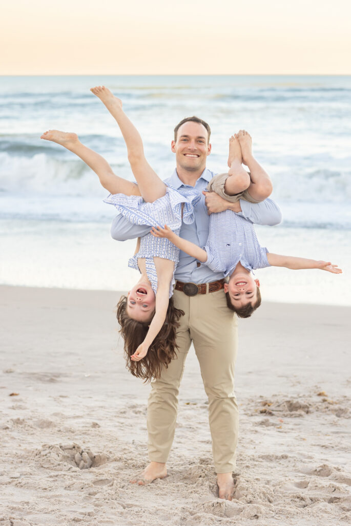 A man stands on a sandy beach, smiling and holding two laughing children upside down, one in each arm. The ocean waves are in the background and the children are wearing light-colored summer clothes. Vero Beach Photoshoot