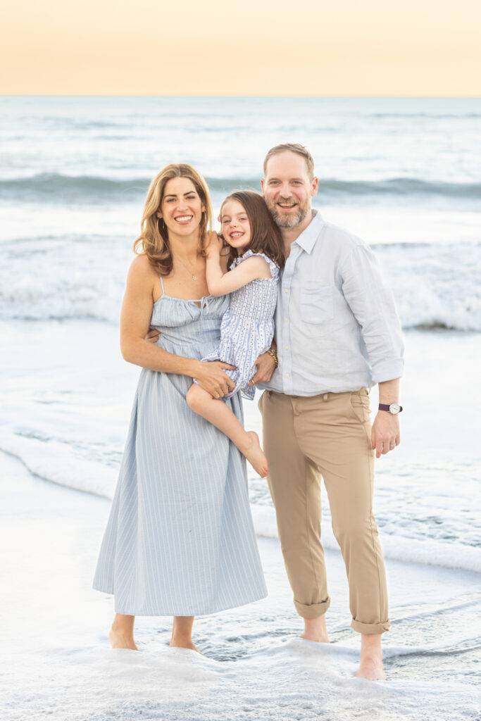 A smiling family of three stands barefoot on the beach. The woman in a blue dress, man in light shirt and tan pants, and child in a striped dress pose together with waves and a sunset in the background. Vero Beach Photoshoot