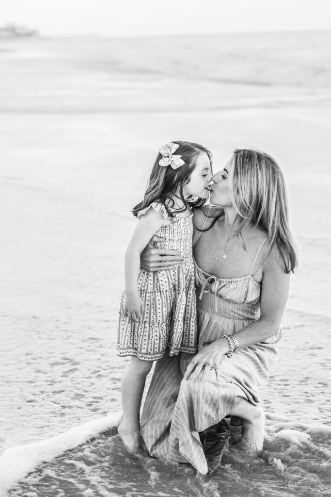 A woman kneels in shallow ocean water, hugging a young girl who kisses her on the nose. Both wear summer dresses. The scene is candid and warm, set on a beach with gentle waves in the background. Vero Beach Photoshoot