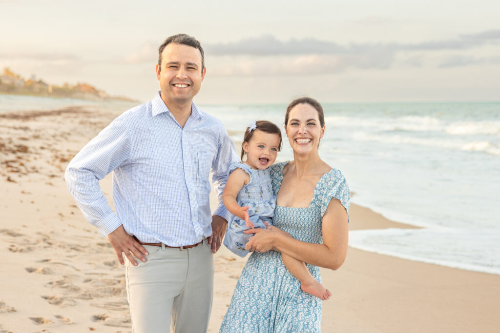 A smiling man, woman, and young child stand together on a sandy beach near the ocean. The woman holds the child, and all three wear light blue clothing. The sky is partly cloudy and the waves are gentle. Vero Beach Photoshoot