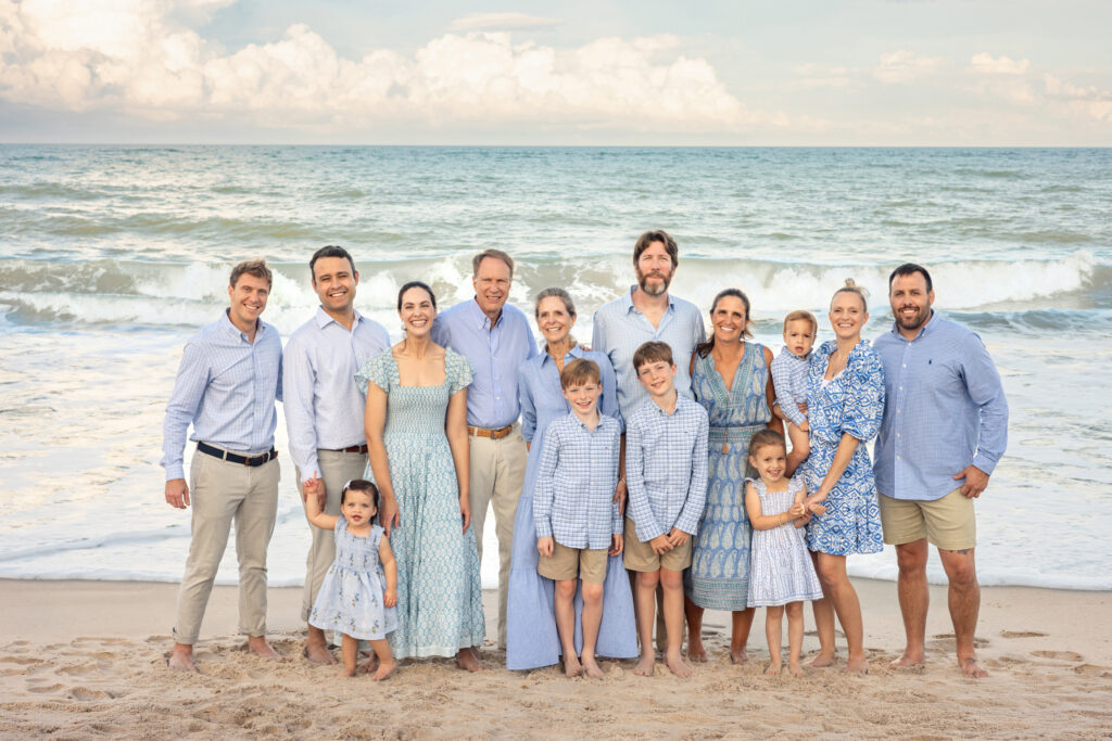 A large family group of fifteen, including adults and children, pose together on a sandy beach with waves and a cloudy sky in the background. Most are dressed in blue and white clothing, smiling at the camera. Vero Beach Photoshoot