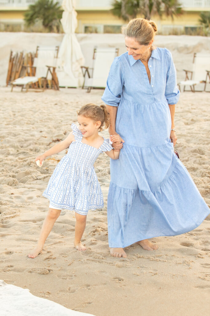 An older woman and a young girl, both in light blue dresses, walk barefoot together on a sandy beach, holding hands and smiling near the shoreline with lounge chairs in the background. Vero Beach Photoshoot