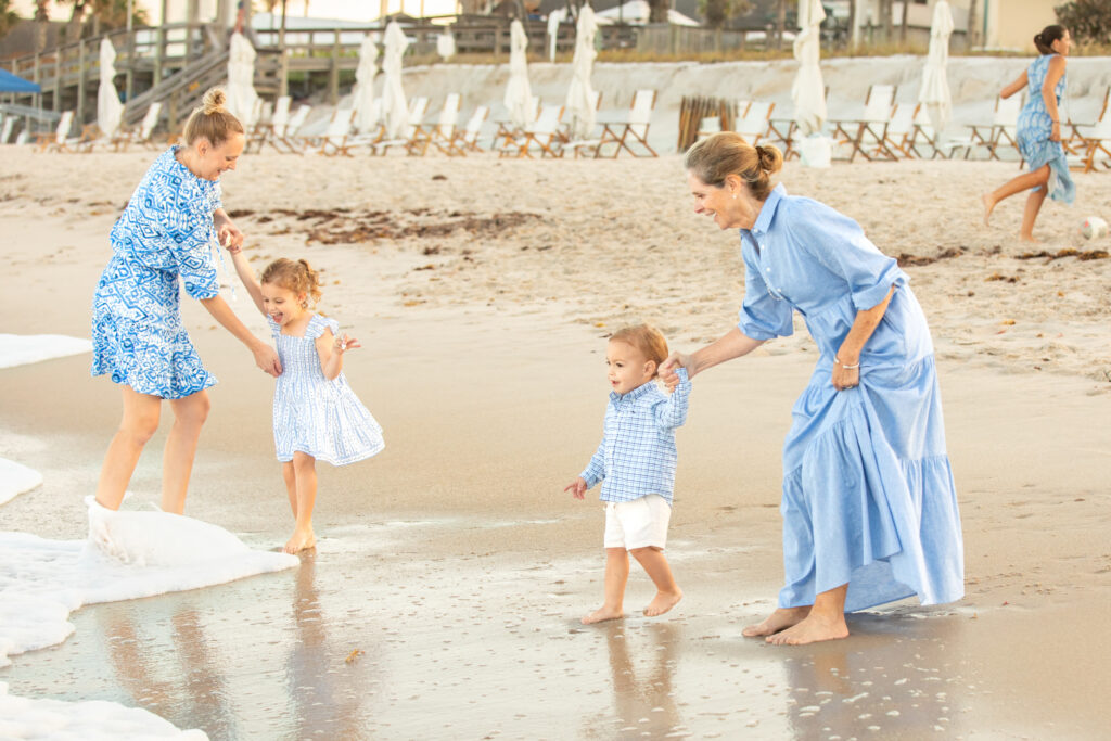 Two women and two young children walk barefoot at the edge of the ocean on a sandy beach. All are wearing blue outfits, and the adults hold the children’s hands while gentle waves wash onto the shore. Vero Beach Photoshoot