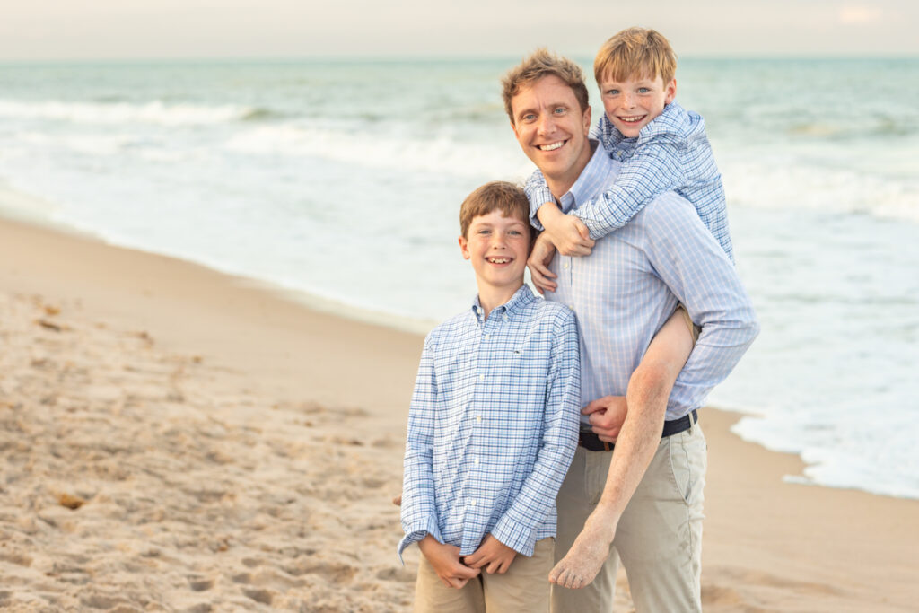 A man stands on a sandy beach with two young boys; one boy stands in front smiling, while the other rides piggyback, both boys and the man wearing blue plaid shirts and smiling at the camera. Waves are in the background. Vero Beach Photoshoot