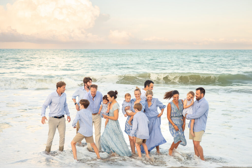 A large family stands together at the beach shoreline, smiling and laughing as waves splash their feet. They wear coordinated blue and white outfits, and the sky is partly cloudy over the calm ocean. Vero Beach Photoshoot
