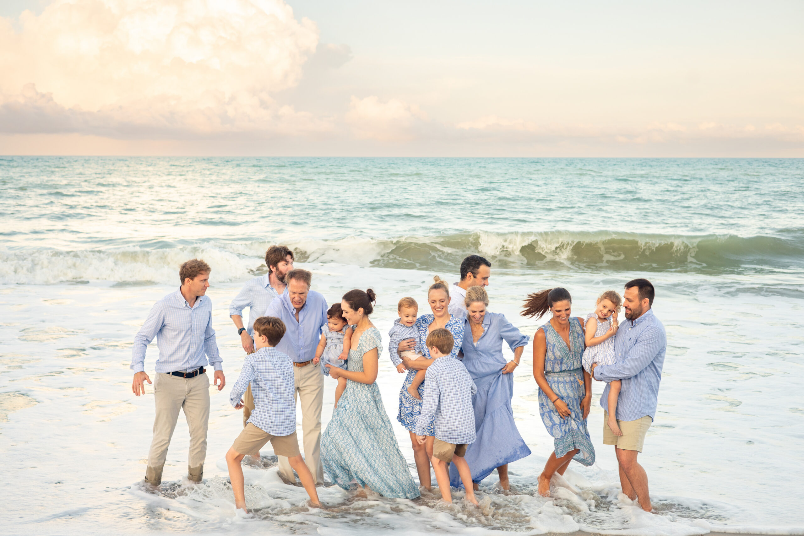 A large family stands together at the beach shoreline, smiling and laughing as waves splash their feet. They wear coordinated blue and white outfits, and the sky is partly cloudy over the calm ocean. Vero Beach Photoshoot