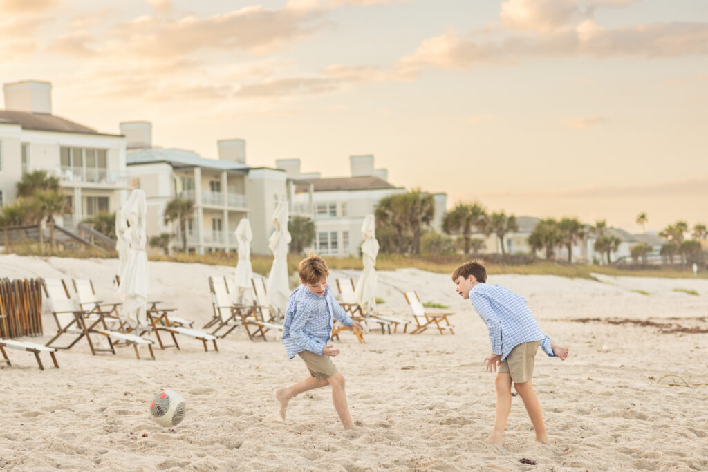 Two young boys wearing blue shirts and khaki shorts play soccer on a sandy beach near vacant lounge chairs and umbrellas, with beachside buildings and palm trees in the background at sunset. Vero Beach Photoshoot