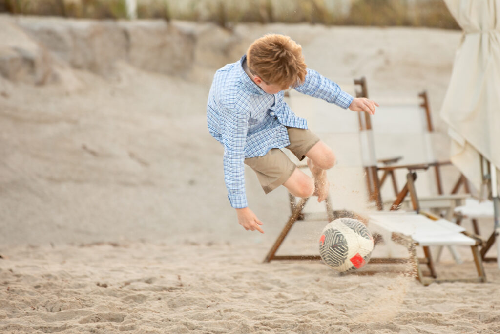A boy wearing a blue plaid shirt and khaki shorts kicks up sand as he jumps to kick a soccer ball on a sandy beach, with deck chairs and a beach umbrella in the background. Vero Beach Photoshoot