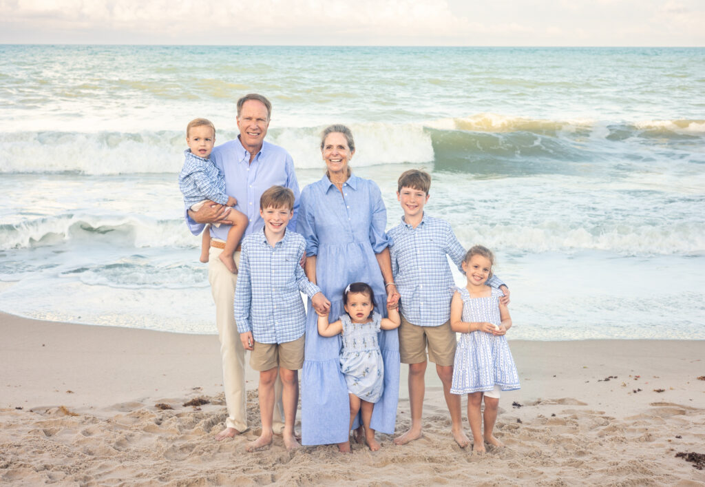 A smiling older couple stands on a sandy beach with ocean waves behind them, surrounded by five young children. Everyone is dressed in coordinated light blue and beige outfits. Vero Beach Photoshoot