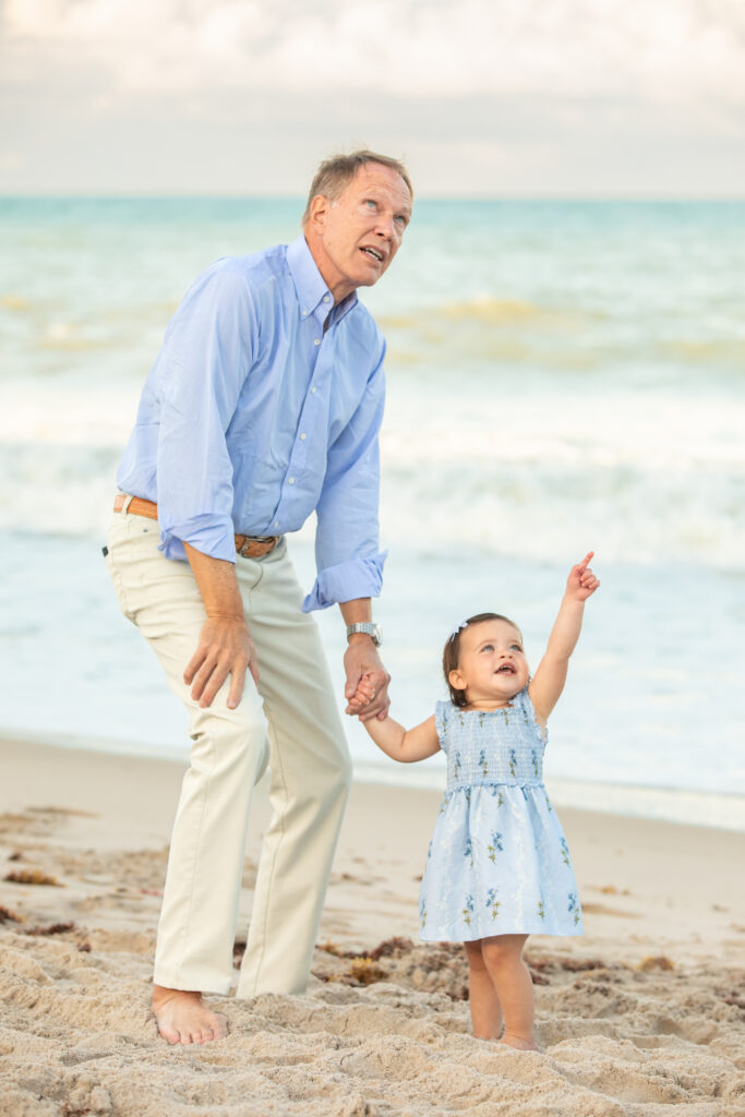 A man and a young girl stand on a sandy beach. The man holds the girls hand and looks up, while the girl points toward the sky and smiles. The ocean waves and cloudy sky are in the background. Vero Beach Photoshoot