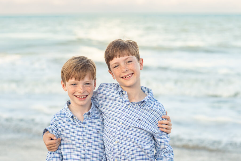 Two young boys with light hair and wearing matching plaid shirts smile with their arms around each other, standing on a sandy beach with gentle waves and the ocean in the background. Vero Beach Photoshoot