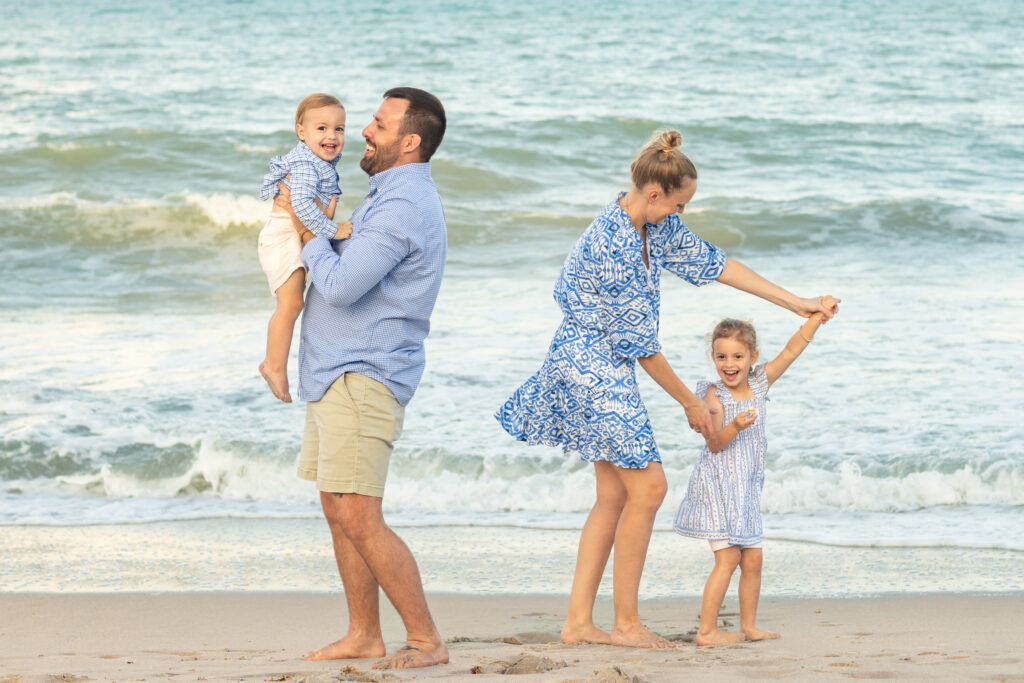 A family of four, dressed in blue and white, smiles and plays together on a sandy beach with waves in the background. The father lifts a toddler while the mother twirls an older child. Vero Beach Photoshoot