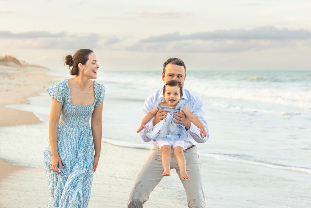 A smiling family of three enjoys time together on a sandy beach. The mother in a blue dress laughs, while the father, in light clothes, holds their happy baby near the shoreline with gentle waves in the background. Vero Beach Photoshoot