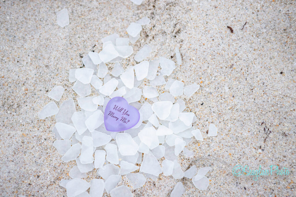 A heart-shaped purple stone with &ldquo;Will You Marry Me?&rdquo; written on it is surrounded by white sea glass pieces, all placed on sandy beach. Vero Beach Photoshoot