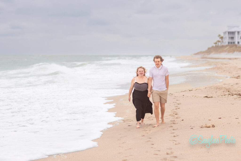 A smiling couple walks hand-in-hand along an empty beach, with gentle waves and cloudy skies in the background. Both are barefoot; the woman wears a black dress and the man wears a white shirt and tan shorts. Vero Beach Photoshoot