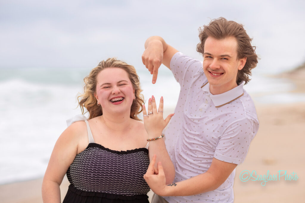 A smiling couple stands on a beach; the woman displays her left hand with a ring while the man beside her points to it, both looking happy and excited. The ocean and sand are visible in the background. Vero Beach Photoshoot