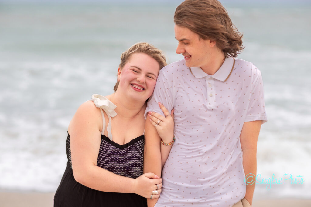 Two people stand close together on a beach, smiling. The person on the left is leaning their head on the person on the rights shoulder. Waves and cloudy sky are visible in the background. Vero Beach Photoshoot