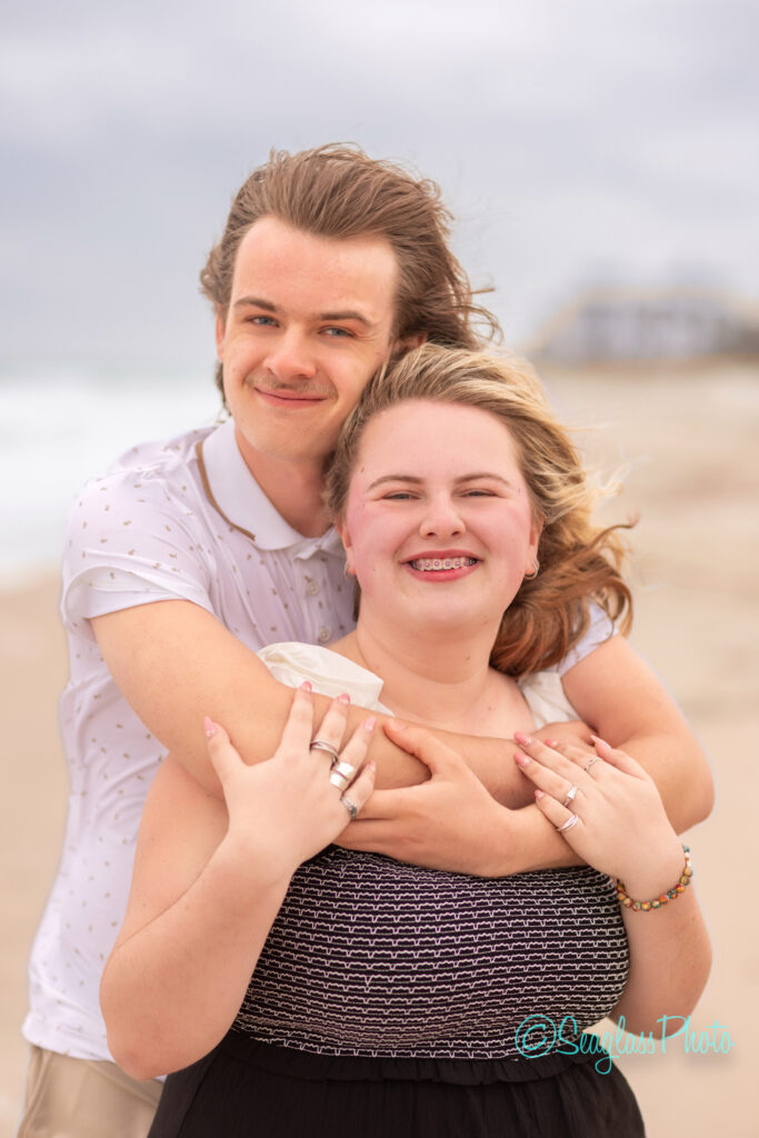 A smiling couple poses on a sandy beach with the ocean in the background. The man stands behind the woman, hugging her with both arms. Both look happy and relaxed, with windblown hair and casual clothing. Vero Beach Photoshoot