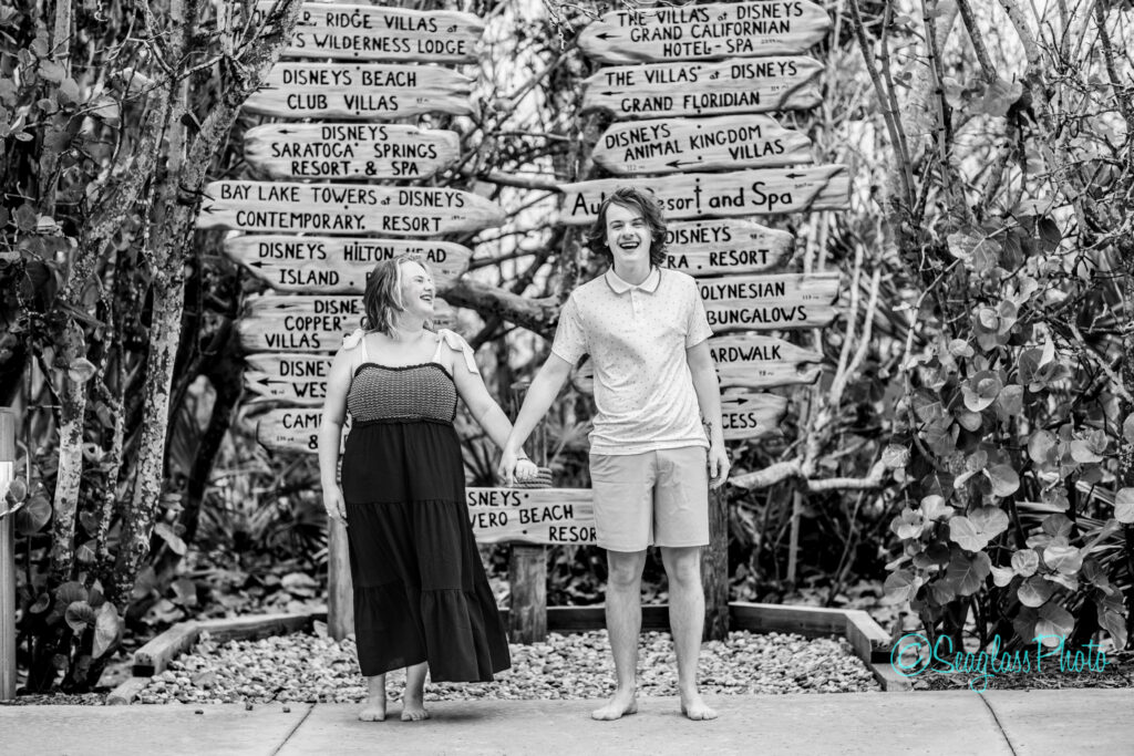 A barefoot couple smiles and holds hands in front of wooden signposts pointing to various Disney resorts, surrounded by lush foliage. The image is black and white. Vero Beach Photoshoot