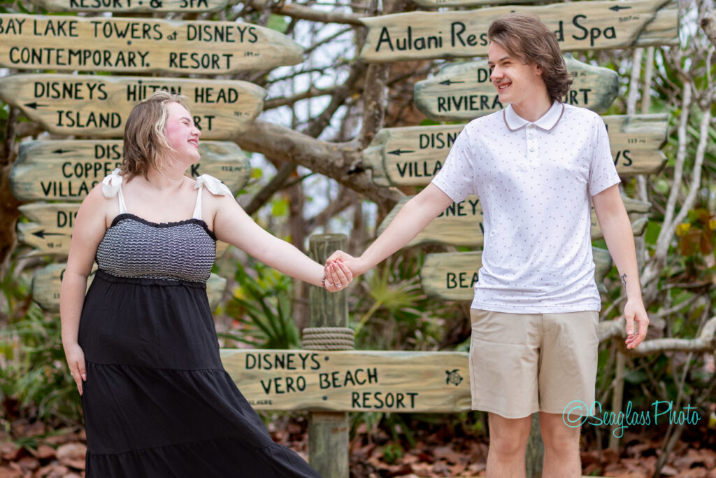 A young couple holds hands and smiles at each other, standing in front of a wooden signpost with multiple Disney resort names. The background is outdoor and leafy, with casual, relaxed vibes. Vero Beach Photoshoot