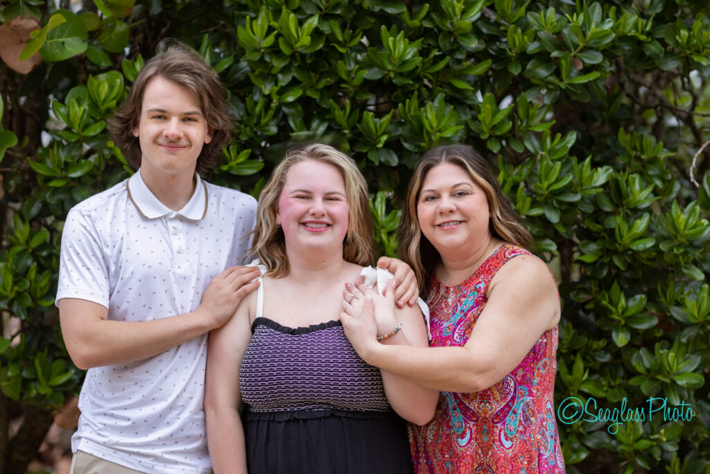 Three people stand close together, smiling in front of leafy green bushes. The person on the left wears a white polo, the center person wears a black dress, and the person on the right wears a colorful sleeveless top. Vero Beach Photoshoot
