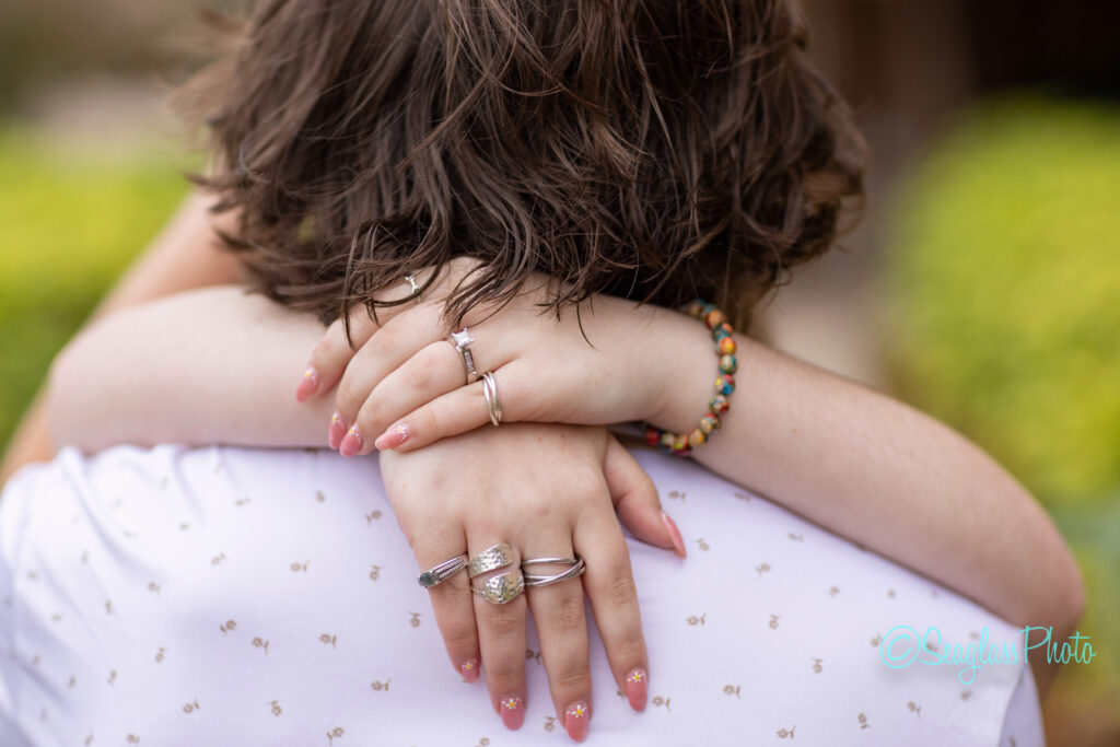 Close-up of a person hugging another, with their hands resting on the others back. The hands have several rings, pink manicured nails with small designs, and a colorful beaded bracelet. The background is blurred greenery. Vero Beach Photoshoot
