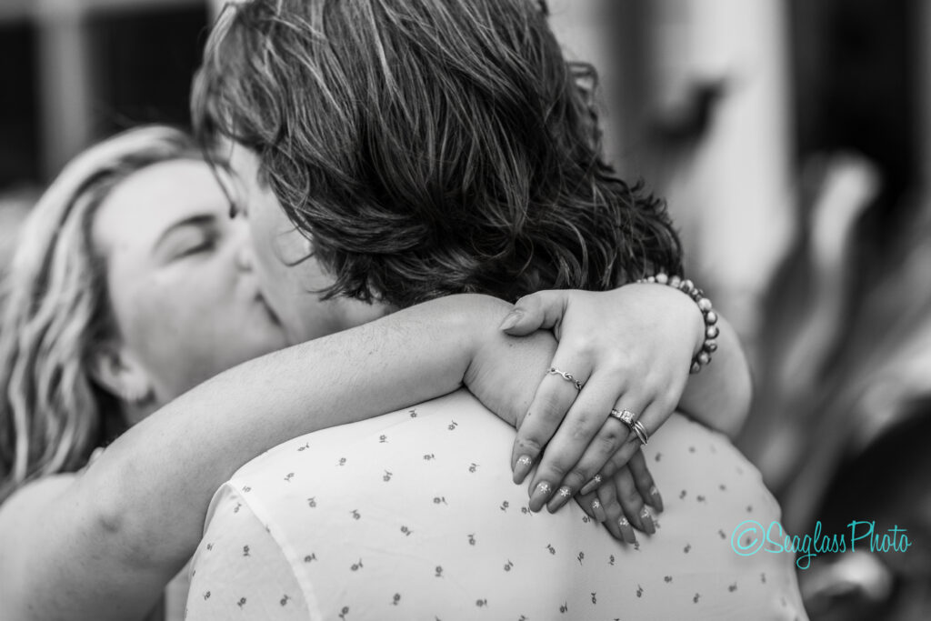 A black and white photo showing two people embracing and kissing, with one person&rsquo;s arms around the other&rsquo;s neck, displaying rings and manicured nails. The focus is on their hands and closeness. Vero Beach Photoshoot