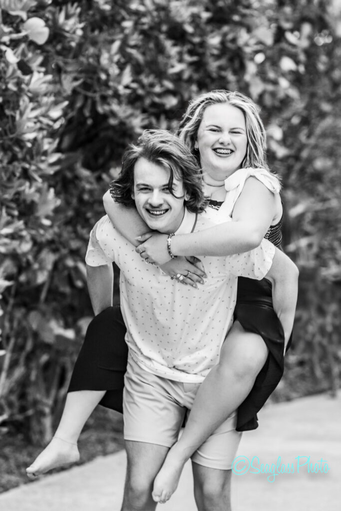 A young man gives a piggyback ride to a smiling young woman on a path lined with leafy bushes. Both are laughing and appear joyful. The image is in black and white. Vero Beach Photoshoot