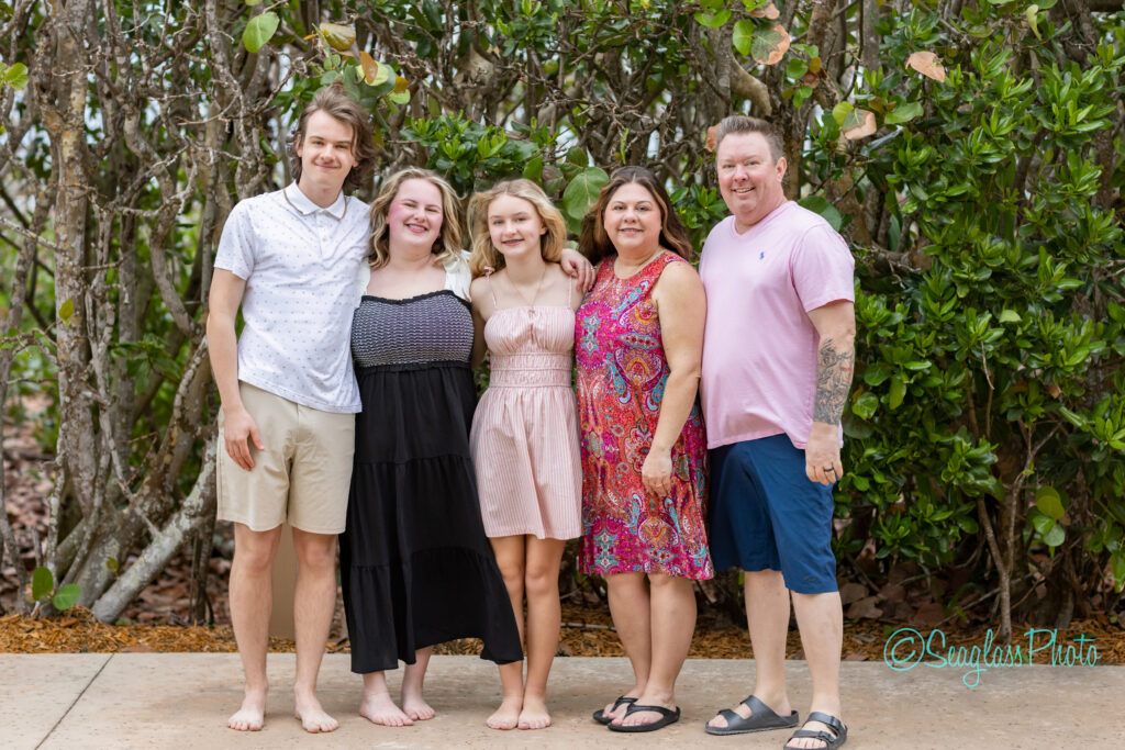 A family of five stands barefoot outdoors, smiling in front of green leafy bushes. The group includes two adults and three children, all dressed in casual summer clothing. Vero Beach Photoshoot