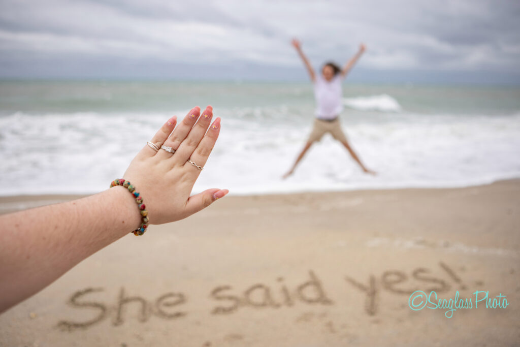 A hand with an engagement ring is held out in focus, while a person in the background jumps with joy on a beach. She said yes! is written in the sand near the waters edge. Vero Beach Photoshoot
