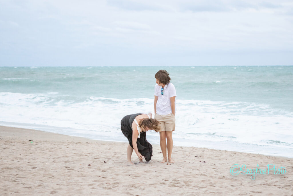 A woman in a black dress bends down on a sandy beach while a man in a white shirt and shorts stands beside her, looking on; waves and cloudy sky are visible in the background. Vero Beach Photoshoot