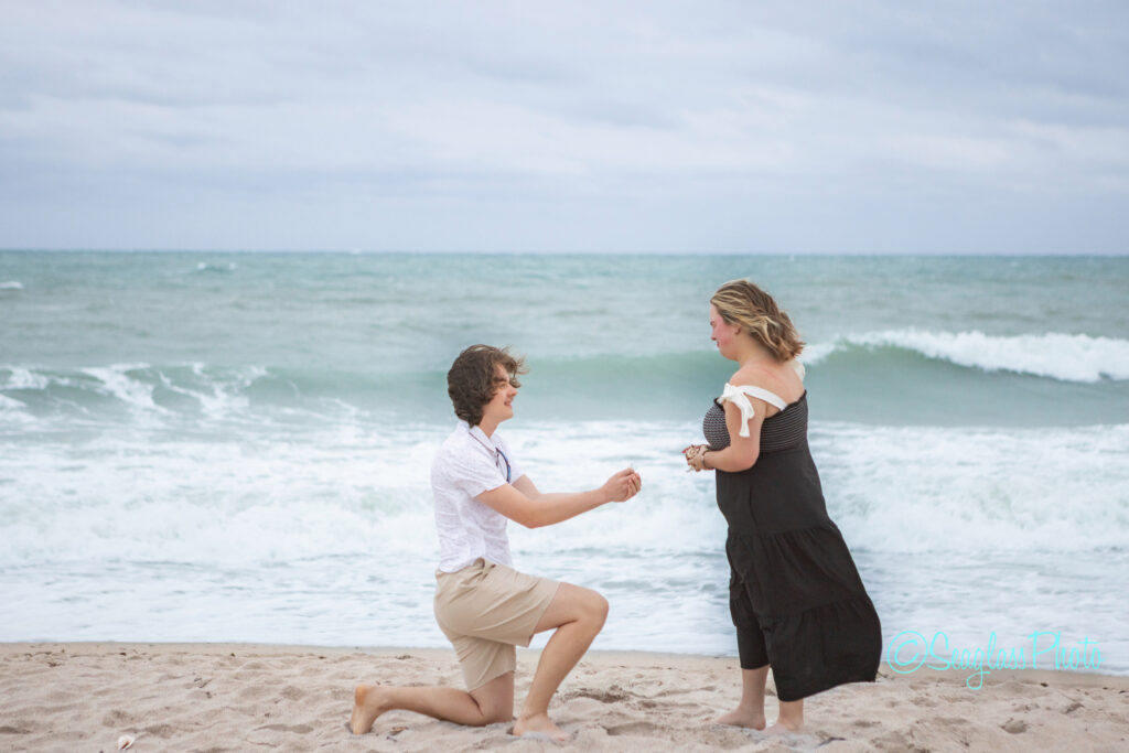 A person kneels on the sand at the beach, holding out a ring toward another person who looks surprised and happy. The ocean waves are in the background under a cloudy sky. Vero Beach Photoshoot