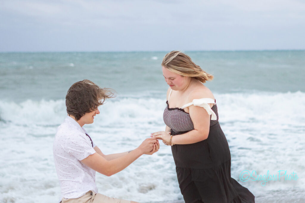A person kneels on the beach, placing a ring on another persons finger as ocean waves crash in the background. Both appear happy and emotional during the proposal moment. Vero Beach Photoshoot