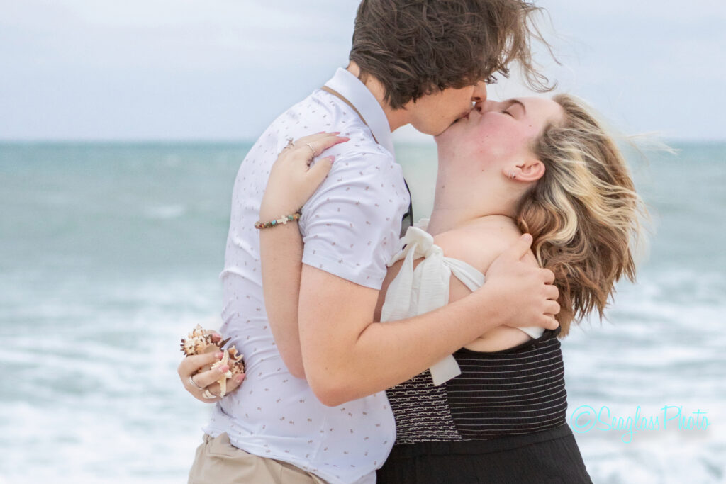 A couple shares a kiss on a beach with waves in the background. One person holds a seashell bouquet and both embrace closely, with windswept hair and casual clothes. Vero Beach Photoshoot