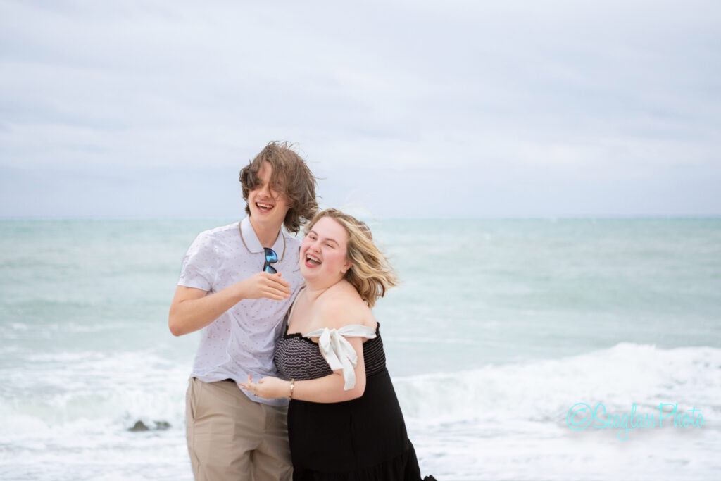 Two people laugh together on a beach, standing close with wind blowing their hair. The ocean and cloudy sky are in the background, creating a joyful and carefree atmosphere. Vero Beach Photoshoot
