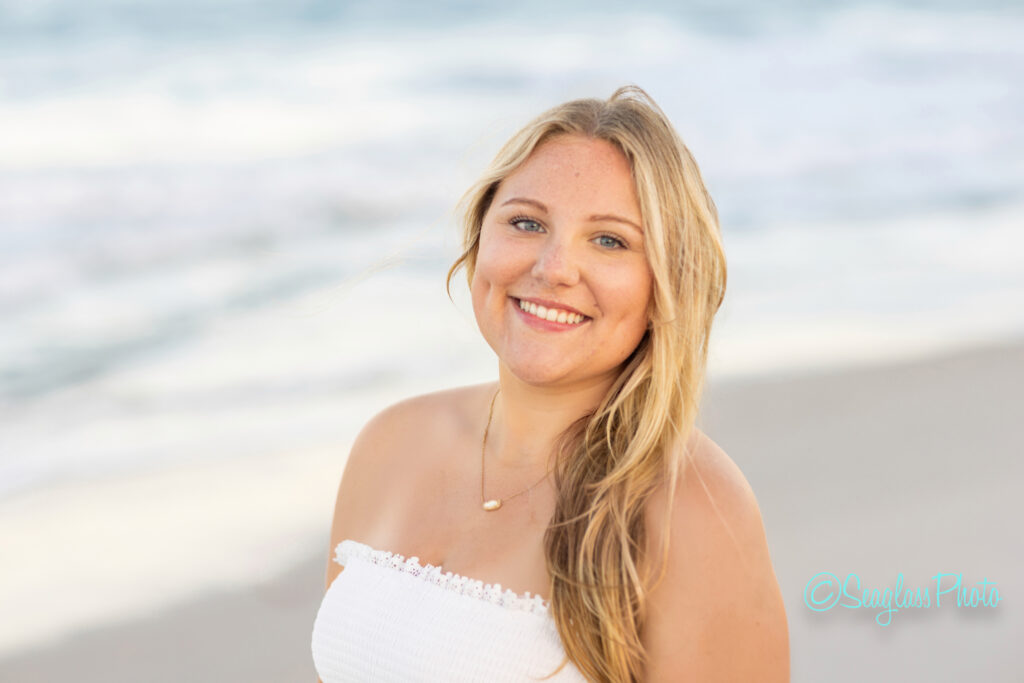 A young woman with long blonde hair and a white strapless top smiles at the camera, standing on a sandy beach with ocean waves in the background. Vero Beach Photoshoot