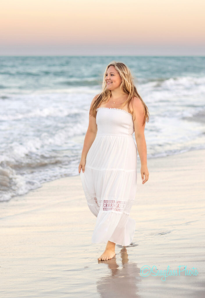 A woman in a long white dress walks barefoot along the beach at sunset, with waves gently touching the shore and a pastel-colored sky in the background. Vero Beach Photoshoot