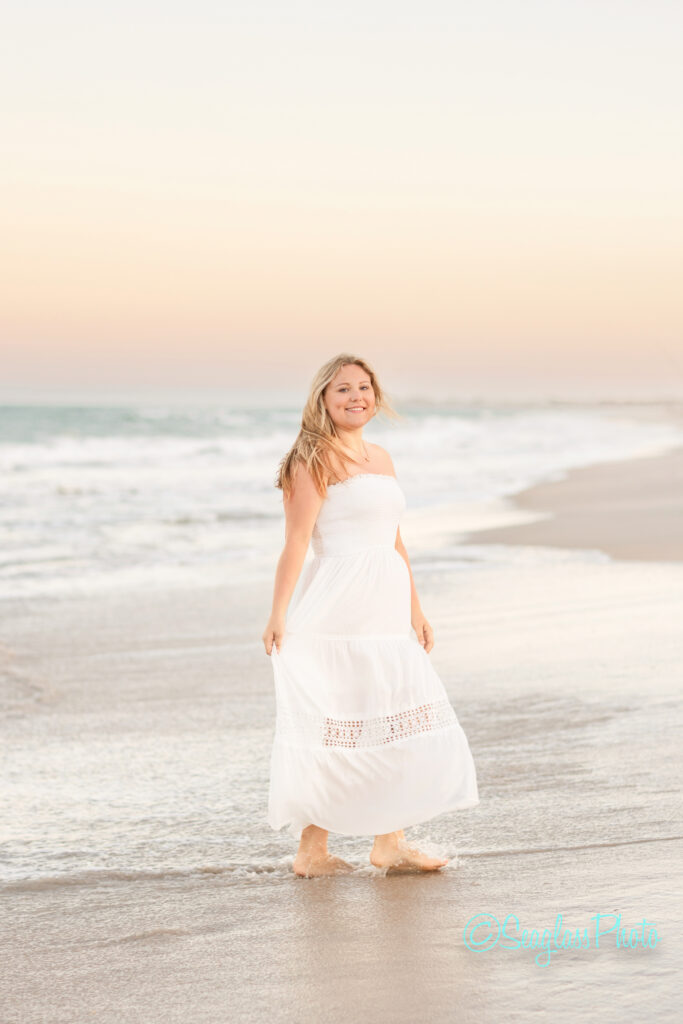 A woman in a white dress walks barefoot along the beach at sunset, smiling with the ocean waves and soft pastel sky in the background. Vero Beach Photoshoot