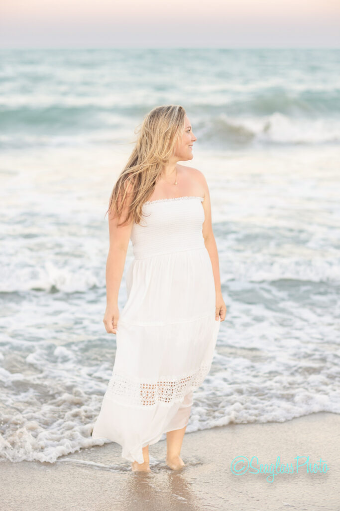 A woman in a white strapless dress stands at the edge of the ocean, looking to the side as gentle waves lap at her feet under a soft pastel sky. Vero Beach Photoshoot
